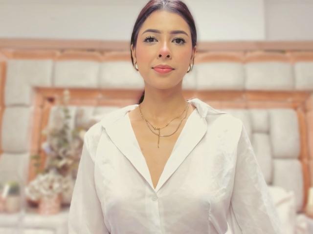 HellenBells has brown hair, tan skin, and wears a white button-up shirt, showing subtle breasts. She has gold earrings and a necklace, smiling softly at the camera.
