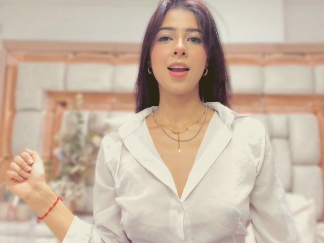 HellenBells with long dark hair, white button-up shirt, gold necklaces, and red bracelet, slightly smiling, holding hand up. Boobs visible under shirt. Blurred background.