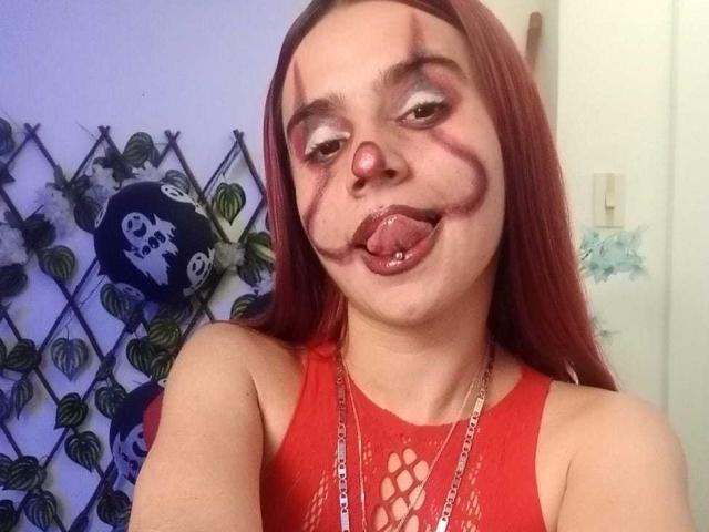 LoveMaxim with red hair, makeup, and nose ring, tongue out, wearing a red mesh top, showing pierced tongue and facial markings, in a selfie.