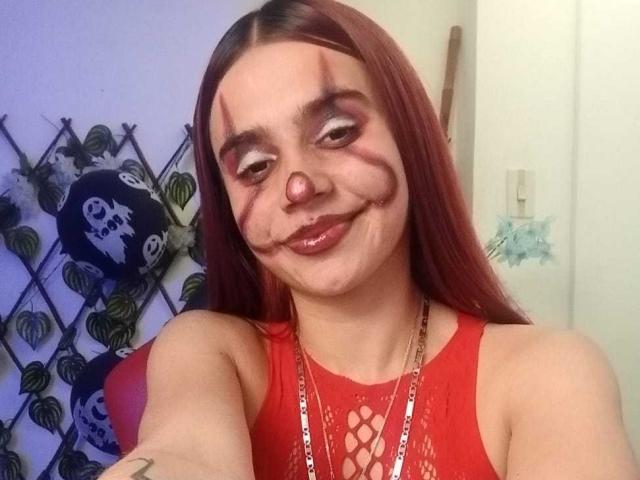 LoveMaxim has red fishnet dress, dark makeup with ghostly face, smiling with nose slightly up, red hair, and a lanyard. Her tits are small, she's cute.