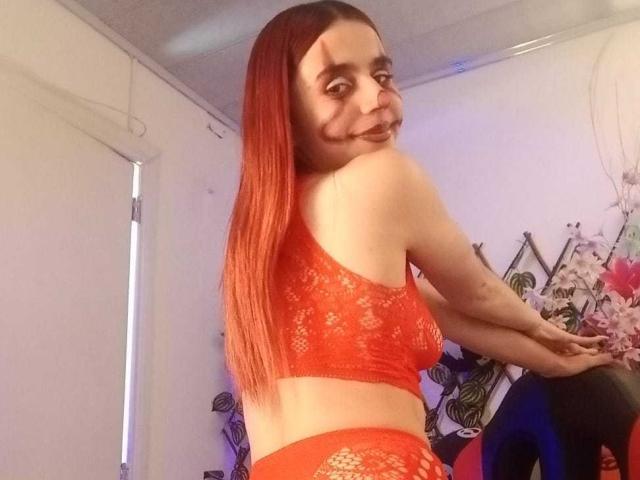 LoveMaxim with red lace bra and panties, facing sideways, showing off her ass, with long red hair, and a playful face with dark makeup. Room has soft lighting.