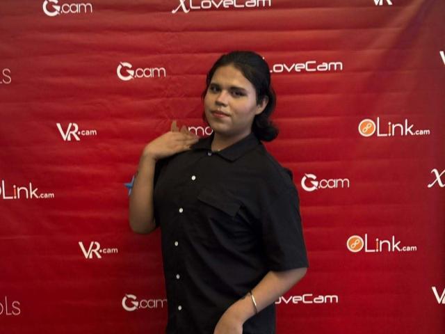 ANGELROUSSE with dark hair in a black shirt, standing, hand near chest, looking at camera, red background, slight smile, wearing a bracelet, medium skin, attractive.