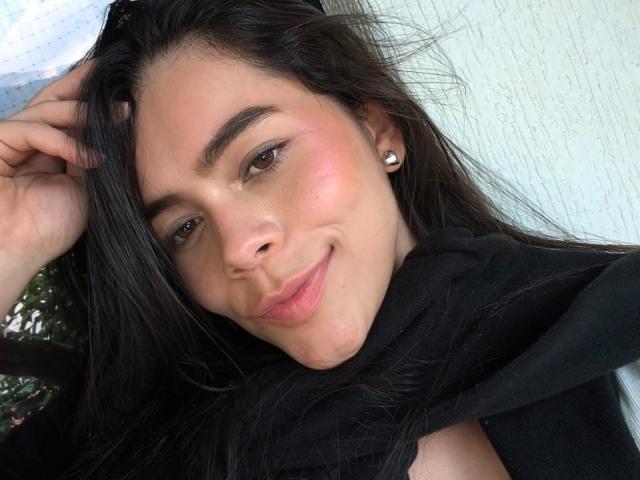 AlanaBlake with long black hair, brown eyes, and a slight smile, wearing a black scarf, and small pearl earrings, showing a subtle blush on her cheeks.