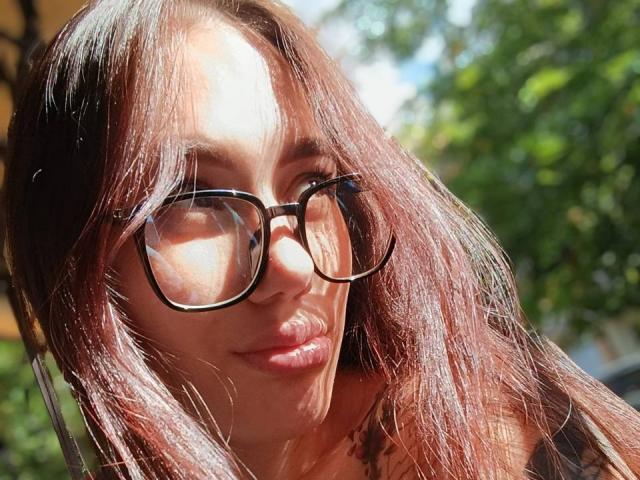 GeraNia with brown wavy hair, wearing black glasses, sunlight on her face, slightly smiling, soft pink lipstick, green blurred background, looking slightly to the right.