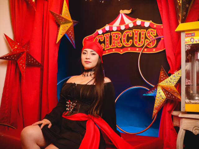Ann Fisher, avec de longs cheveux bruns, portant un foulard rouge, une robe noire à épaules dénudées avec un ruban rouge, et un collier noir, assise devant un décor sur le thème du cirque avec des étoiles..