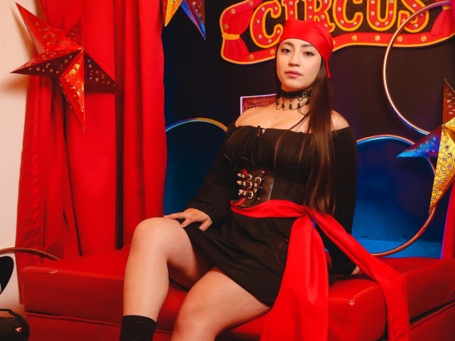 AnnFisher with long brown hair, wearing a red headband, black off-shoulder dress, choker, and red ribbon, sitting on red bench, showing bare thighs, with a star decoration.