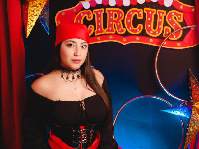 Ann Fisher mit langen, braunen Haaren, die einen roten Hut, ein schwarzes Oberteil mit einem Schulterfrei-Schnitt, eine schwarze Halskette und ein rotes Korsett trägt, zeigt ihre Brüste, in einer Zirkus-Themen-Kulisse mit bunten Dekorationen..