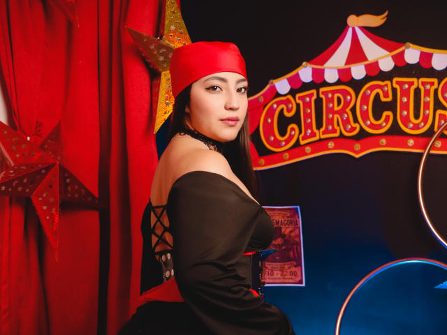 Ann Fisher mit roter Mütze, schwarzem, schulterfreiem Kleid, Schnürung im Rücken, die ihr Gesäß zeigt, dunklen Augen, Make-up, sitzend, Zirkus-Thema im Hintergrund, Stern, roter Vorhang, Ring am rechten Handgelenk..