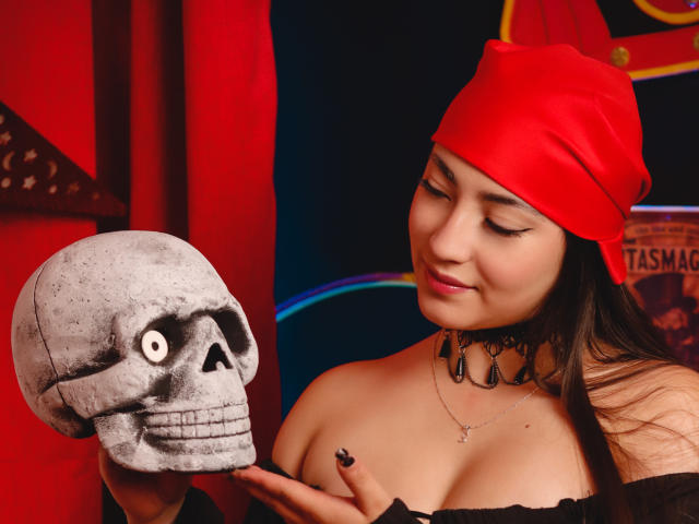 Ann Fisher, portant un bonnet rouge, un collier noir et un haut à épaules dénudées, tient un crâne aux grands yeux, sourit, montre ses seins et porte du vernis à ongles noir..