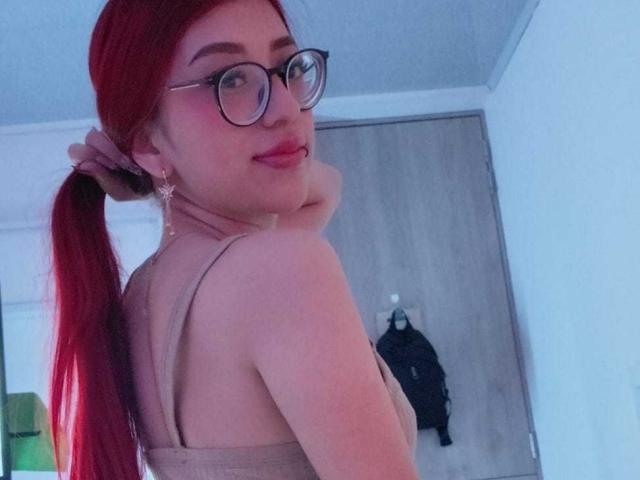 CherryBuchel with red pigtails, glasses, and pink lipstick, showing off her bare back and tan top, looking over her shoulder with a slight smile. Her tits are small.
