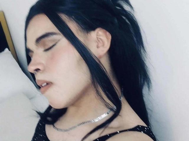 ANGELROUSSE with black hair, eyes closed, soft pink blush, slightly open mouth, silver necklace, black top, leaning against white pillow, looking relaxed and sleepy.