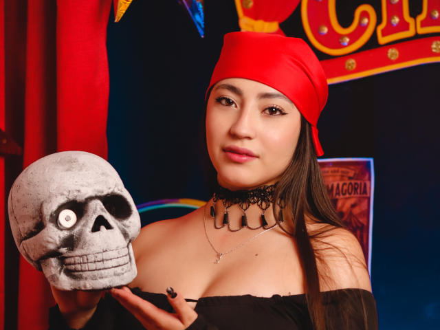 AnnFisher with long brown hair, wearing a red headscarf, black choker, and off-shoulder top, holding a skull, showing her small tits and a confident smile.