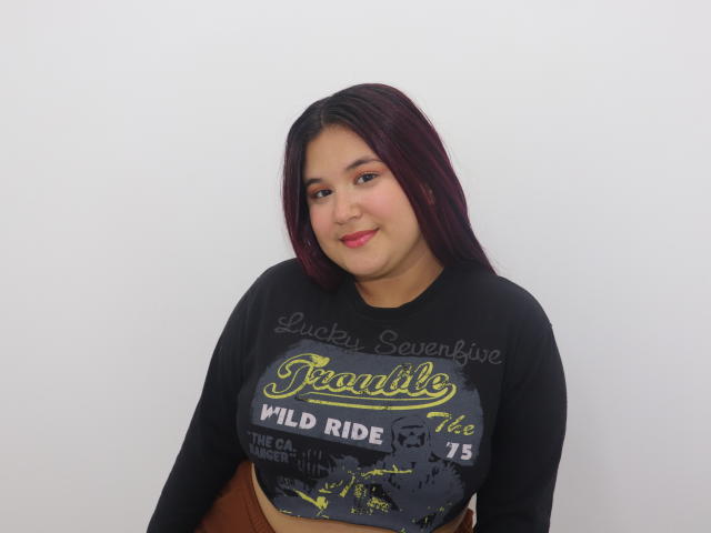 KityMiss with dark purple hair, black crop top showing tits, smiling at camera, brown pants, white background, watermark "Luckysavenise" across chest.