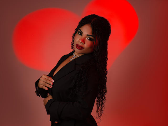 NahiaraReina with curly black hair, red lipstick, black blazer, and necklace, stands against a red heart, looking over her shoulder, hands near her chest. Boobs visible under blazer.