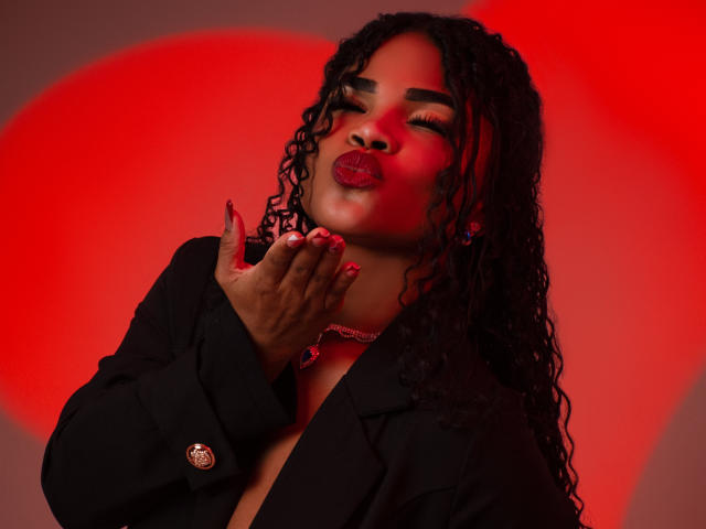 NahiaraReina with curly black hair, red lipstick, and black blazer, pouting with hands near face, red glowing background, wearing choker and pink nail polish, looking sexy.