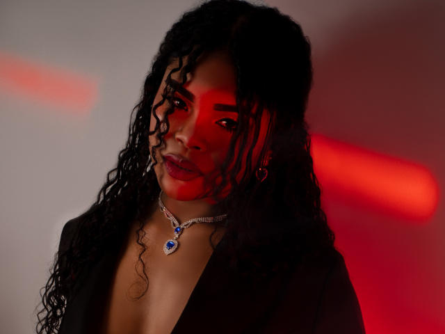 NahiaraReina with curly black hair, red-lit face, deep cleavage, wearing a black top and silver necklace with a blue gem. Red light casts dramatic shadows.