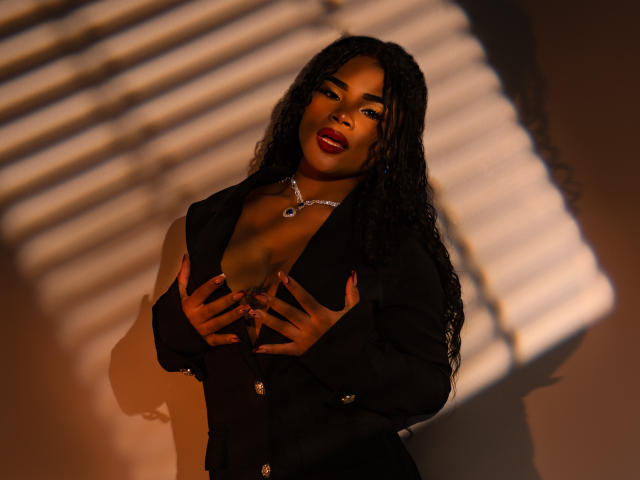NahiaraReina with dark skin, long curly hair, red lipstick, black blazer, hands covering her big, round tits, wearing a silver necklace, standing in warm, shadowy light.