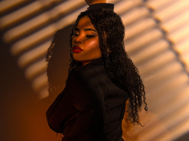 NahiaraReina with curly black hair, red lipstick, and a black top, looking over her shoulder, showcasing her tits, shadowed by warm light and striped patterns.