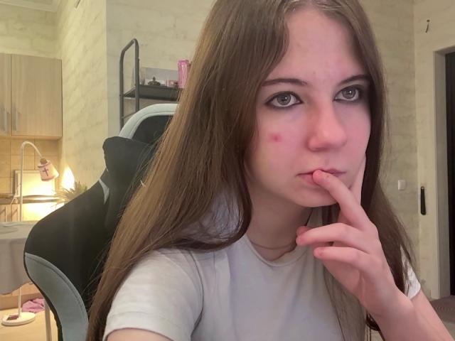 GeorgeCorpeno with long brown hair, pouty expression, finger in mouth, white shirt, slight red blemish on cheek, sitting in black and white gaming chair.