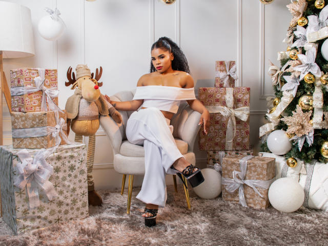 NahiaraReina with curly hair, white off-shoulder top, and white pants, sitting on a chair, holding a reindeer toy, surrounded by Christmas gifts and a decorated tree.