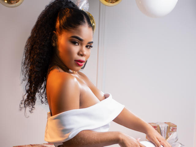 NahiaraReina with curly black hair, red lipstick, and white off-shoulder top, showing her small, perky tits, sitting with arms on a table, looking at the camera.
