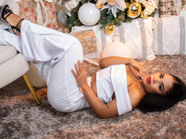 NahiaraReina with curly black hair, laying on a furry rug, wearing an off-shoulder white dress, showing her ass, with black strappy heels, surrounded by Christmas decorations.