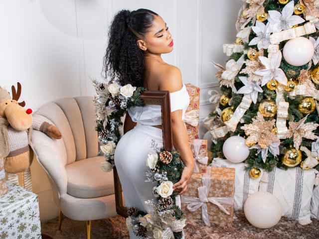 NahiaraReina with curly black hair, red lipstick, white off-shoulder top, and white pants, holding a frame, standing by a Christmas tree with gold and white ornaments. Boobs, ass.
