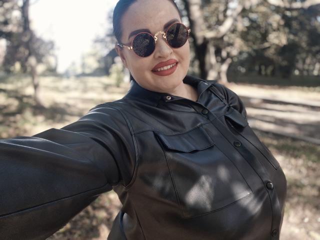 GeorgiaLerox, con rossetto rosso scuro, occhiali da sole rotondi e una camicia di pelle nera, sorride, allungando un braccio per un selfie, mostrando il suo seno prosperoso e un'aria sicura in un parco soleggiato..