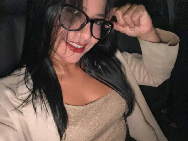 AlanaLuxe, avec ses cheveux noirs, son rouge à lèvres rouge, ses lunettes, sa veste beige et son haut scintillant, montrant sa poitrine, souriant et tenant des lunettes près de son visage..