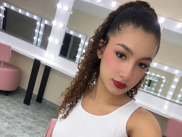 Nathacha Evans tem cabelos castanhos cacheados presos em um rabo de cavalo, veste uma camiseta branca, com blush rosa e batom vermelho, e está em pé em frente a espelhos iluminados em um quarto..