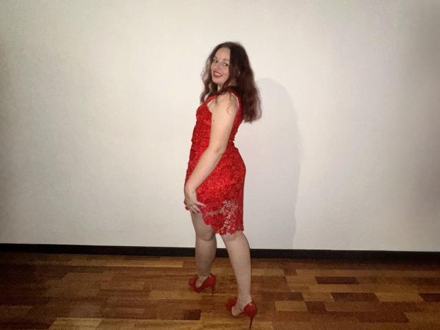 MelodiLady with red lace dress, red high heels, and wavy brown hair, smiling, standing on wooden floor, hands on hips, showing her ass, with pale skin and red lipstick.