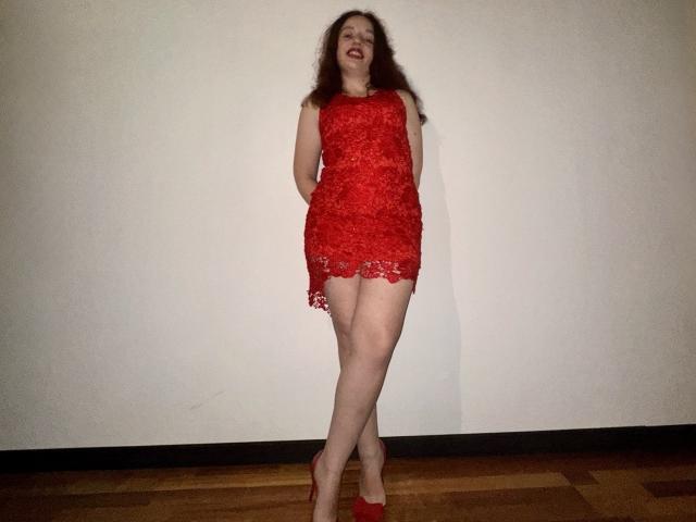 MelodiLady stands in a red lace dress, showing her tits, with her hands behind her back, wearing red heels, smiling, and leaning against a white wall.