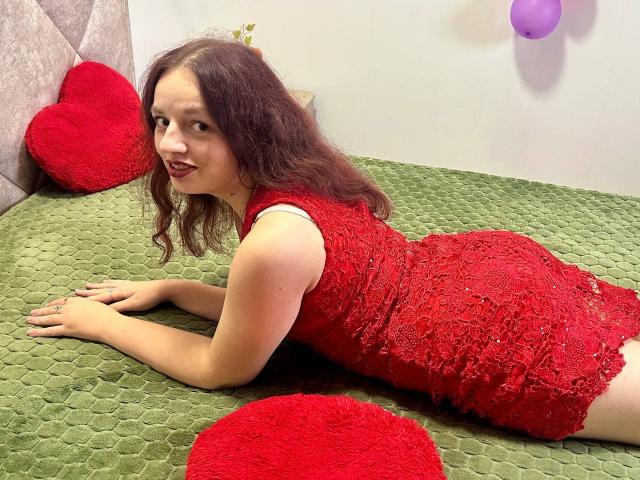 MelodiLady with red lace dress, laying on green bed, showing her ass, red heart pillows, curly brown hair, smiling, ring on left hand, purple wall decoration.