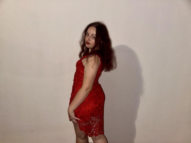 MelodiLady in a red lace dress, standing sideways, showing her ass, with wavy brown hair, red lipstick, and a shadow on a plain wall.