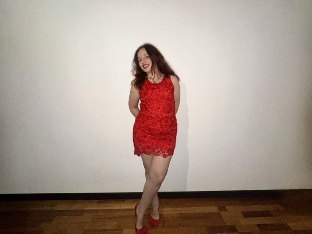 MelodiLady with long brown hair, wearing a red lace dress and red heels, standing with hands behind her back, smiling at the camera, showing her ass and tits.