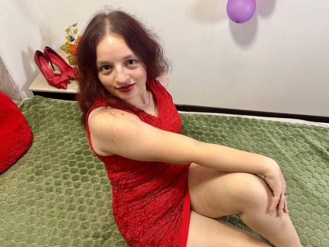 MelodiLady with red lace dress, sitting on green bed, legs crossed, showing her ass, red heels on left, purple balloon on right, wavy brown hair, smiling, red lipstick.