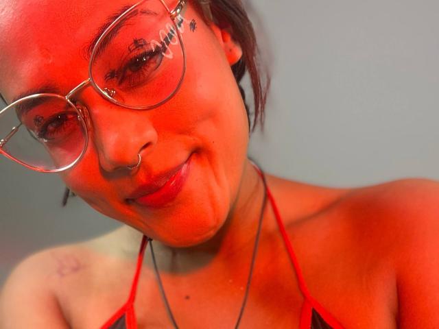 LilithRussoo, con el rostro iluminado en rojo, lleva gafas, un piercing en la nariz y un top halter rojo. Está sonriendo ligeramente, mostrando un pequeño moretón en el hombro izquierdo. Tiene unos pechos pequeños y lindos..