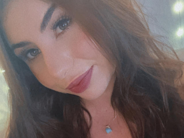 VenusxMusa, avec ses cheveux bruns ondulés, ses sourcils épais, ses yeux bleus et son rouge à lèvres rose, souriant légèrement, portant un collier avec un pendentif bleu, dans un autoportrait avec un éclairage doux..