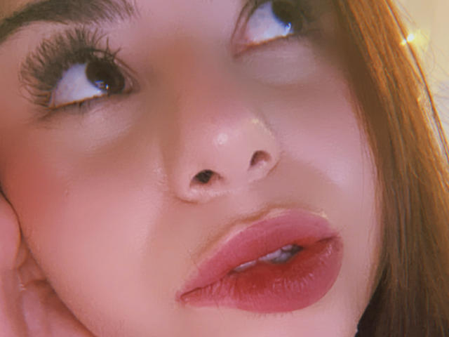Le visage de VenusxMusa, en gros plan, montre des lèvres roses et pulpeuses, légèrement entrouvertes, des yeux marron regardant vers le haut, de longs cils, et une peau rose pâle avec un éclat doux. Ses cheveux roux sont partiellement visibles..