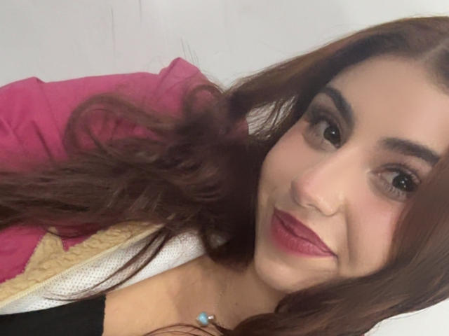VenusxMusa, avec des cheveux bruns, des sourcils épais et du rouge à lèvres, souriant à la caméra, portant un haut rose et jaune, une chemise noire et un collier bleu..