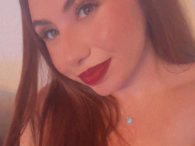 VenusxMusa has red lipstick, blue eyes, and long brown hair. She's wearing a small blue necklace with a gem. Her tits are not visible. She's smiling softly.