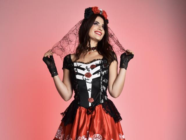 MariaScott with dark hair, wearing a black and red corset with roses, black lace gloves, and a spiderweb veil, smiling, holding her veil, pink background.