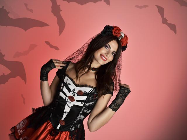 MariaScott with dark brown hair, red roses in hair, black lace gloves, white and black corset with red roses, red satin skirt, black lace veil, smiling, pink background.