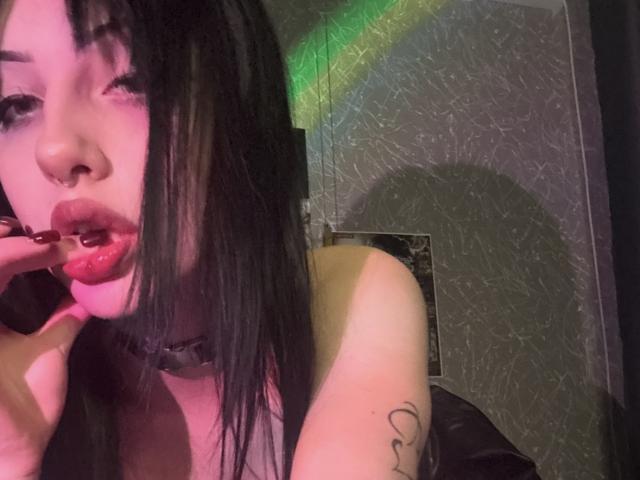 Simonne Brustkern, con cabello negro, piercing en la nariz y lápiz labial rojo, mordiéndose el dedo, mostrando un tatuaje en el brazo superior, usando un collar, en una habitación con poca luz y luz verde..
