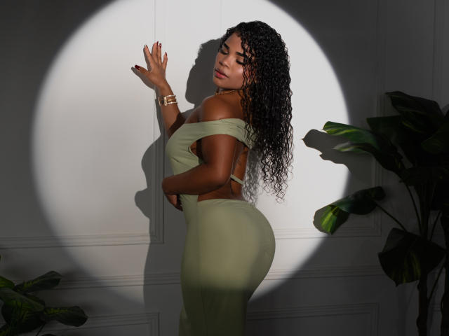 NahiaraReina with curly brown hair, off-shoulder green dress, and tan skin, standing against a wall with a shadow, showing her ass and back, wearing a gold wristwatch.