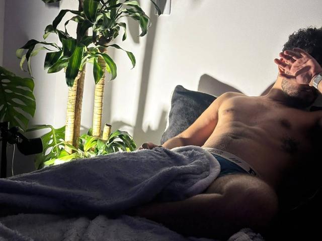CapryWilliams, shirtless with a curly Afro, lies in bed, covered by a gray blanket, wearing blue underwear, hand covering his mouth, surrounded by green plants, in dim lighting.