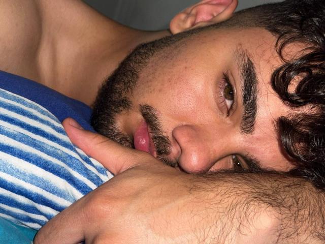 CapryWilliams with curly brown hair, beard, and brown eyes, lying on blue and white striped pillow, finger in mouth, looking at camera, showing soft, hairy arm.