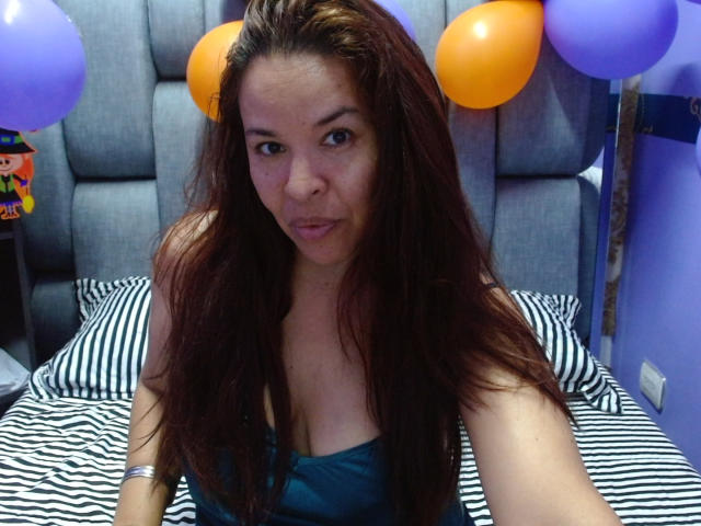 LizethBells has long brown hair, small tits, and a cute face with freckles, wearing a blue top, sitting on a striped bed with orange and purple balloons.