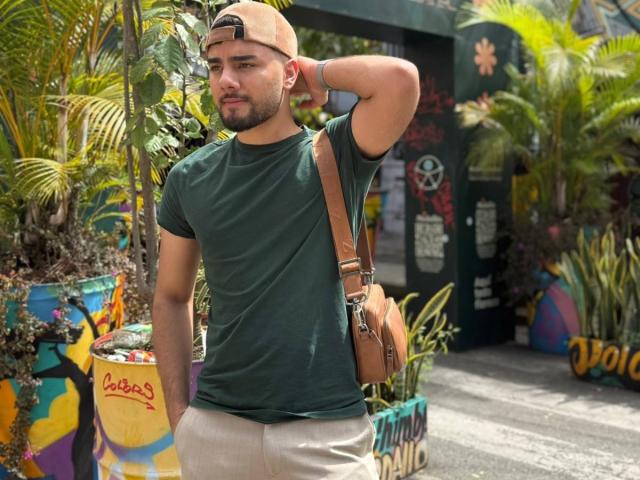 CapryWilliams, a medium-build dude with a green tee, beige pants, brown bag, and tan cap, stands with arm behind head, looking slightly annoyed. His ass is somewhat visible.