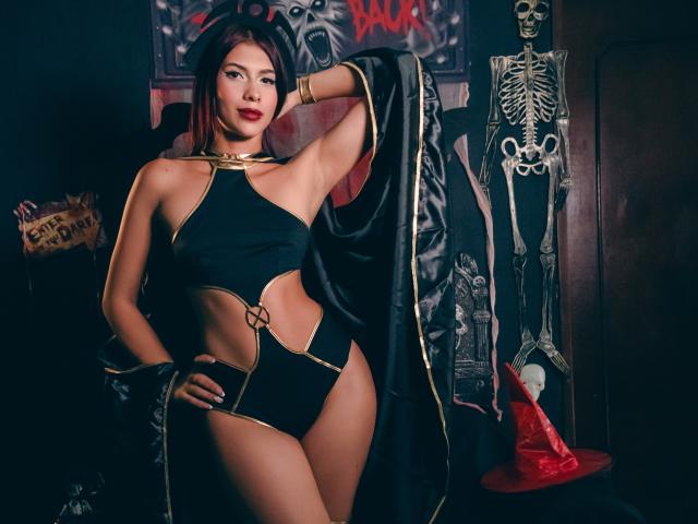 DanikaSerov with dark red hair, wearing a black, gold-trimmed, cutout bikini and black cape, standing with one hand on her hip, showing her small tits and ass.